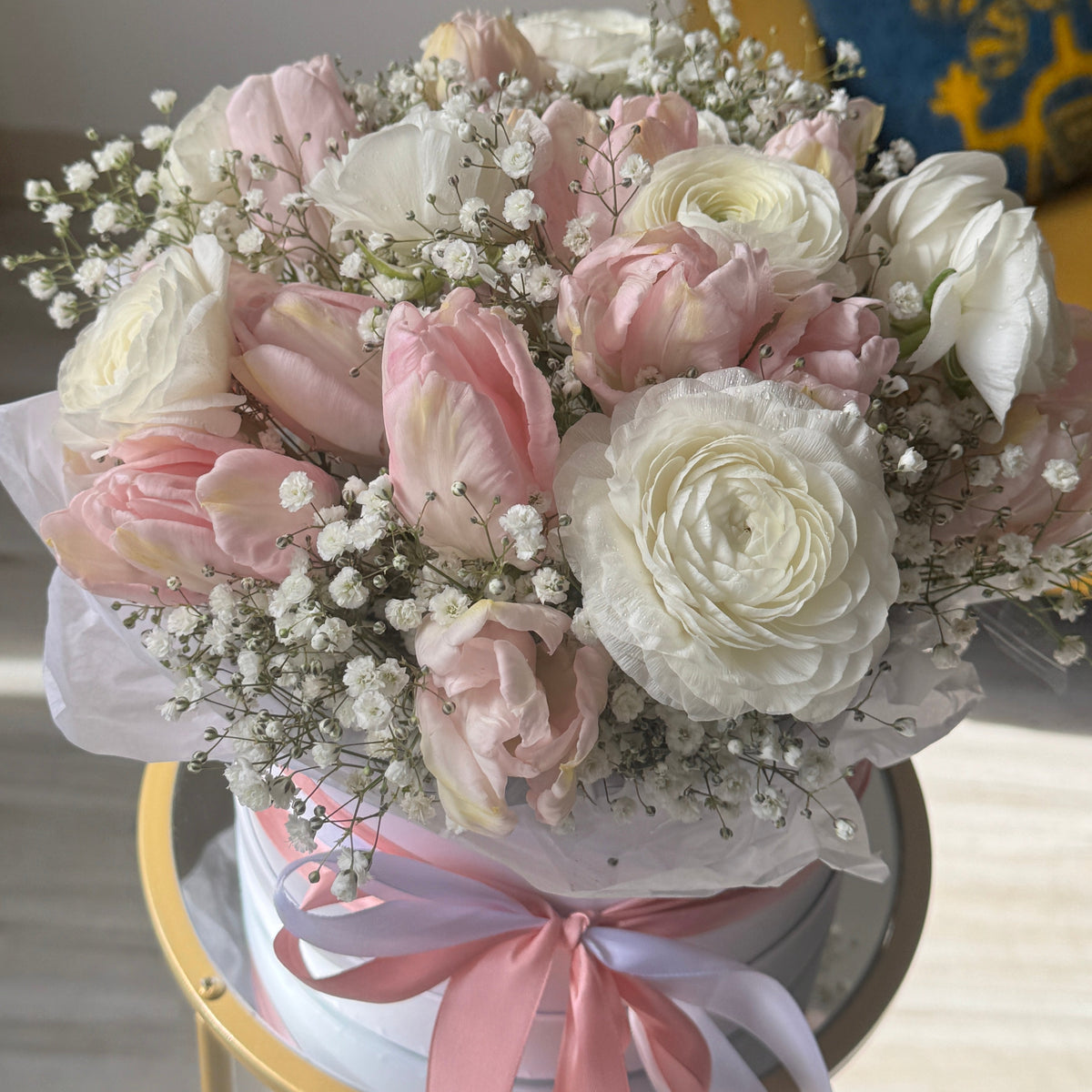 A round floral arrangement of pink and white roses, tulips, and baby's breath in a white box with a pink ribbon, displayed on a gold table in a bright indoor setting.
