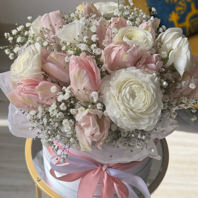 A round floral arrangement of pink and white roses, tulips, and baby's breath in a white box with a pink ribbon, displayed on a gold table in a bright indoor setting.