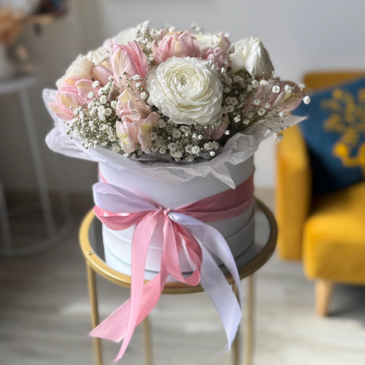 A floral arrangement of pink and white roses with baby's breath in a round white box, tied with a pink ribbon, displayed on a small gold table in a softly blurred indoor setting.