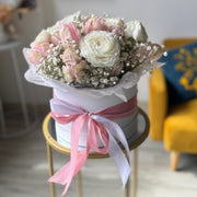 A floral arrangement of pink and white roses with baby's breath in a round white box, tied with a pink ribbon, displayed on a small gold table in a softly blurred indoor setting.