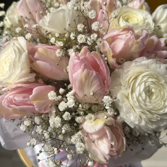 A close-up of a floral bouquet featuring light pink tulips, white roses, and delicate white baby's breath, wrapped in pale pink and white paper with a pink ribbon.