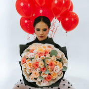 A bouquet with light pink, coral, and white roses in black and white wrapping paper in the hands of a girl with balloons