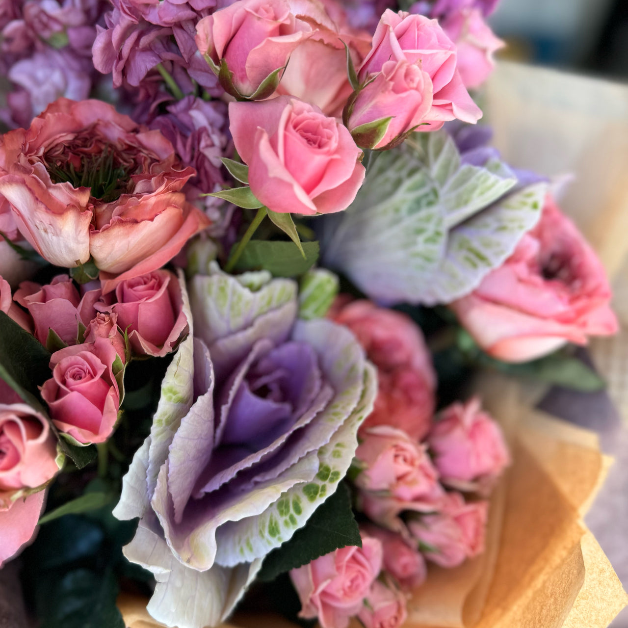 Close-up of the  pink 'Sunset Charm Bouquet,' highlighting the bold purple kale as the focal point. The bouquet features Coral Expression roses, pink spray roses, and mauve stock flowers arranged around the vibrant kale. 