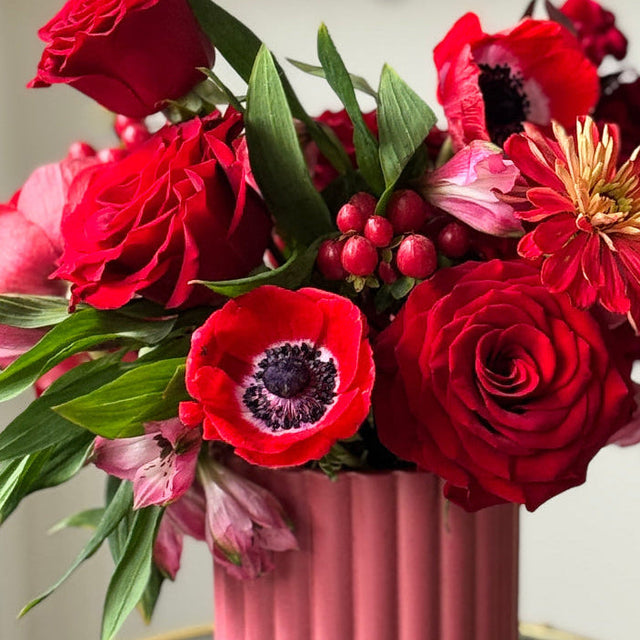 Luxury red floral arrangement in a pink clay vase featuring red roses, anemones, hypericum berries, and seasonal flowers.