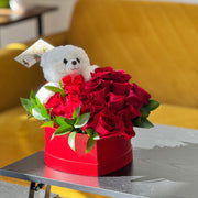 Red box with red roses and a white teddy bear on a table.