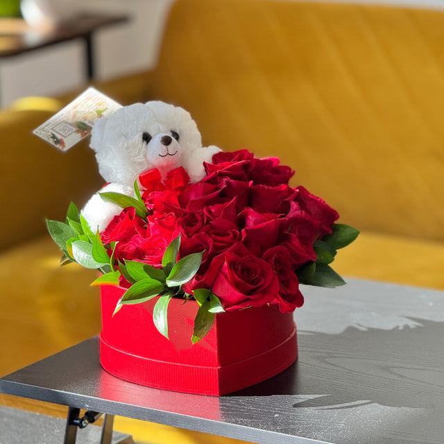 Red box with red roses and a white teddy bear on a table.