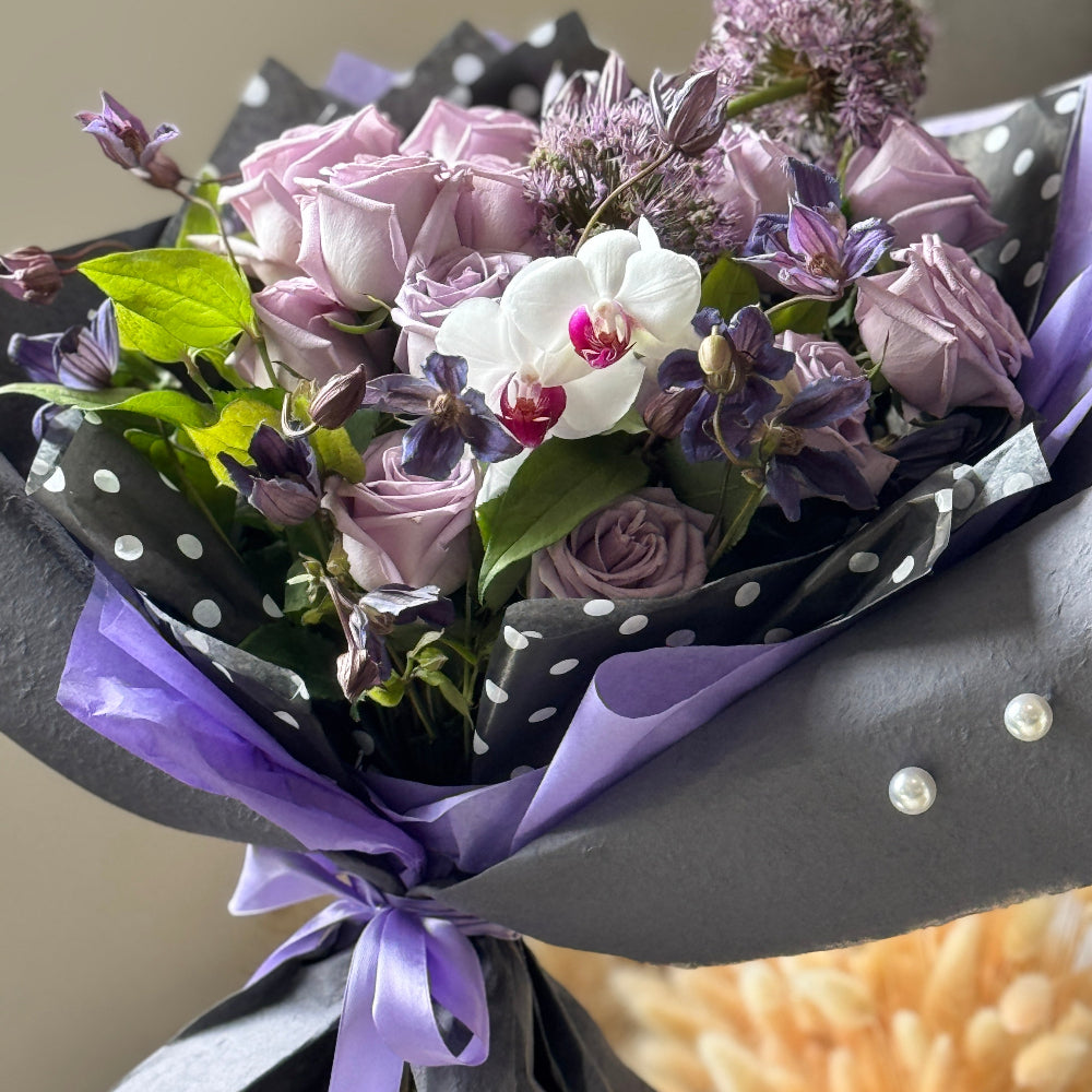 Romantic hand-tied bouquet featuring lavender roses, white orchids, seasonal fillers, and textured greenery in gray wrapping.