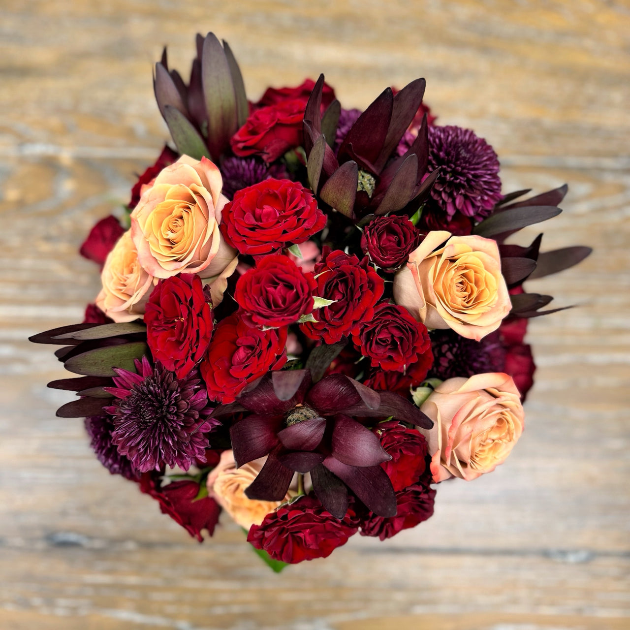 Top-down view of a fall round bouquet  showcasing red spray roses, deep purple mums, cappuccino roses, and orange carnations arranged in a cozy autumn display.