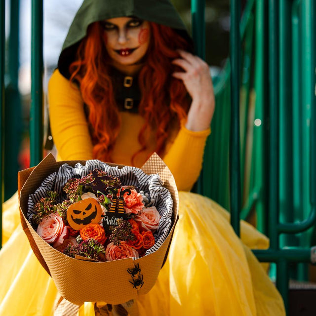 A spooky girl in costume holds a bouquet of orange flowers with scary pumpkins and spiders.