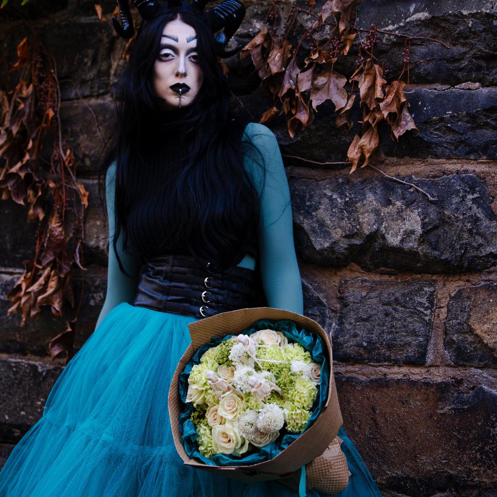 Woman in scary halloween costume is holding the bouquet of white flowers with skeletons.