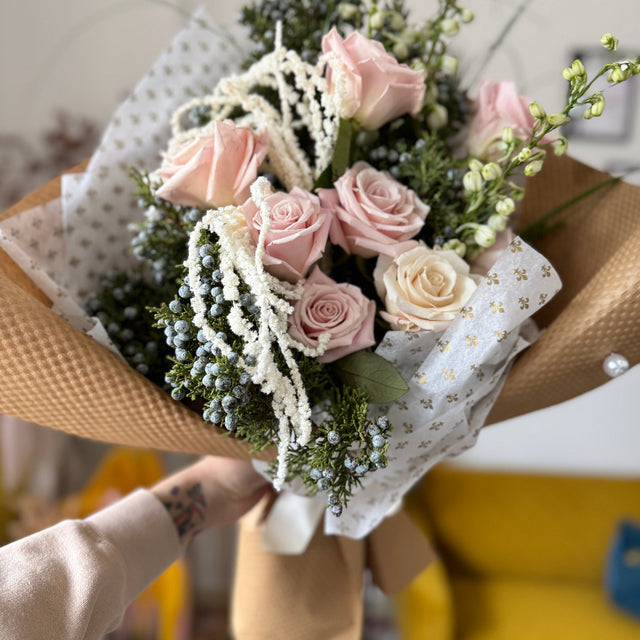 Soft winter bouquet with pink roses, juniper branches, and white delphinium wrapped in kraft paper.