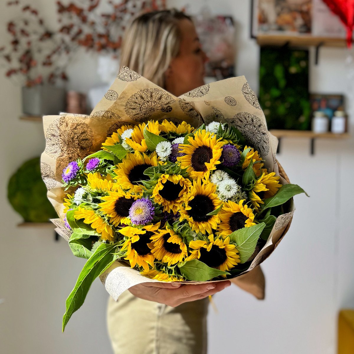 Florist holding a vibrant bouquet featuring sunflowers and purple and white asters. The bouquet is wrapped in eco-friendly brown tissue and kraft paper.