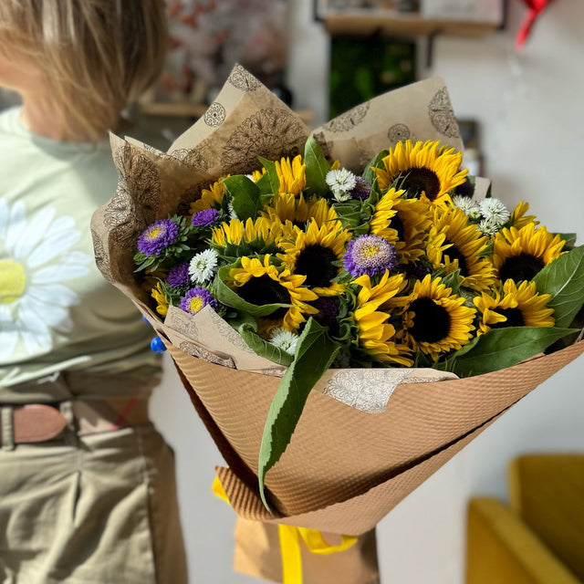 Florist holding a vibrant bouquet of sunflowers and asters wrapped in eco-friendly brown tissue and kraft paper, showcasing a cheerful garden-fresh look."