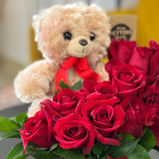 Teddy bear with a red bow sitting behind a bouquet of red roses on a table.