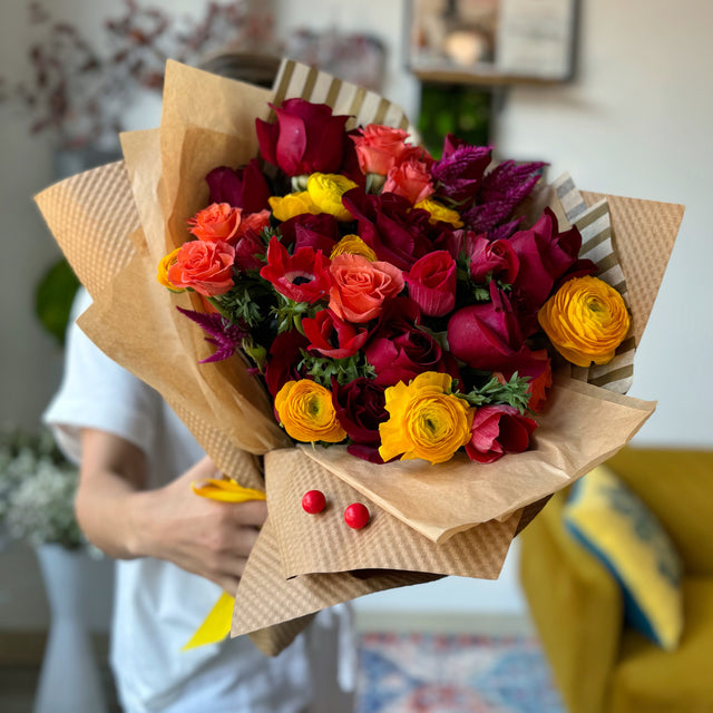 Juicy Fall bouquet wrapped in kraft paper, held by a florist, showcasing vibrant yellow ranunculuses, red roses, orange spray roses, and red anemones.