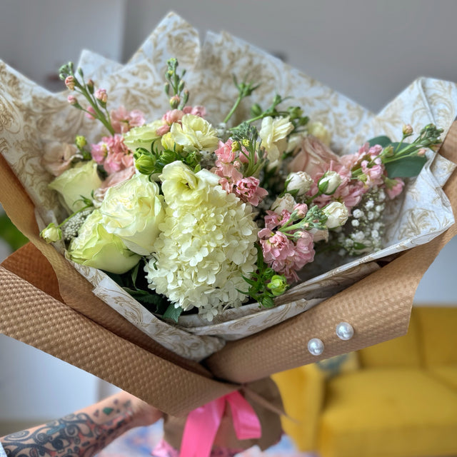 Elegant bouquet featuring white hydrangeas, white roses, ranunculus, and light pink stock flowers, wrapped in white tissue with a gold pattern.