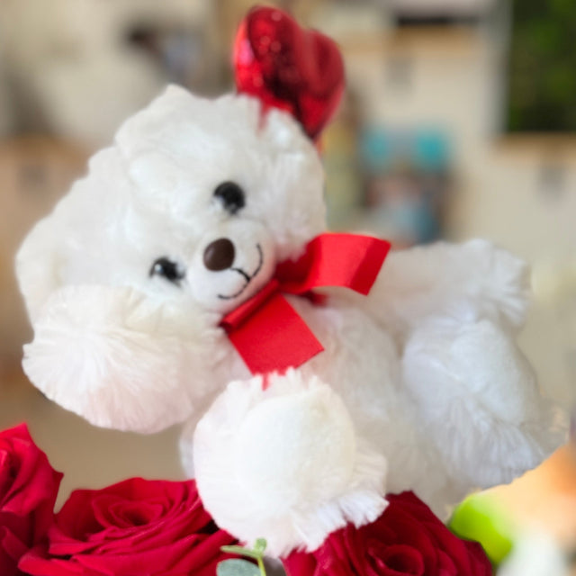 White teddy bear with a red bow surrounded by red roses on a blurred background