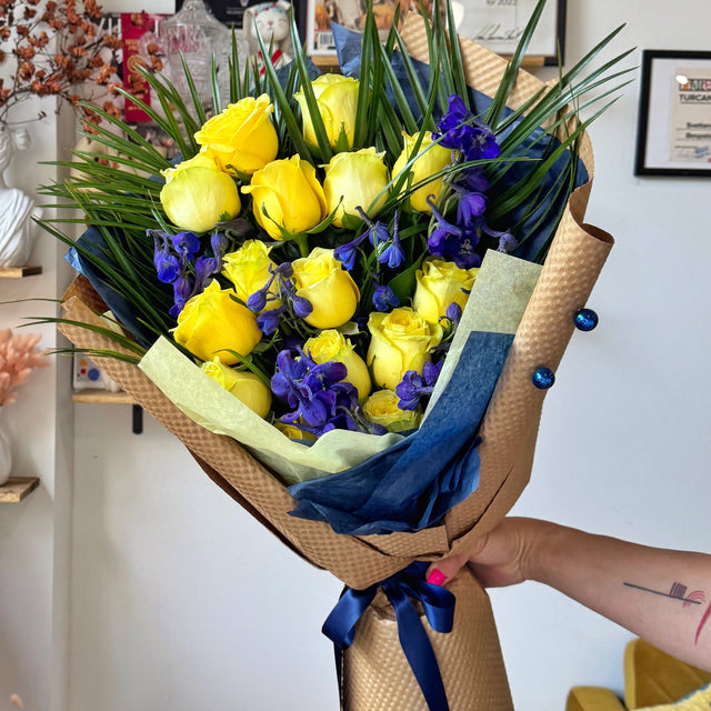  Graduation bouquet featuring yellow roses, blue delphinium, and palm leaves