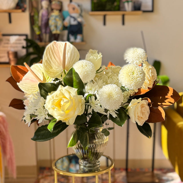 Winter Grace arrangement featuring white roses, hydrangeas, mums, and anthuriums with glossy magnolia leaves in a modern vase.