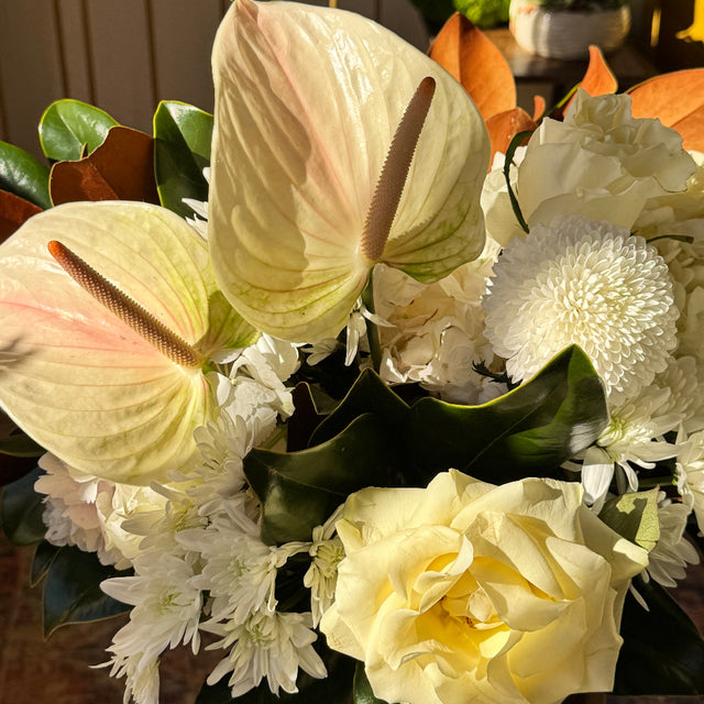 Bouquet of flowers with white and green tones, including roses and anthuriums, in a blurred indoor setting.