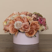 Side view of a floral arrangement in a light pink hat box, featuring cream roses, terracotta hydrangeas, and light pink lisianthus. The arrangement presents a stylish and elegant display with earthy tones.