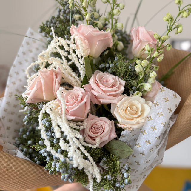 Elegant floral bouquet with pastel roses, winter greens, and hanging amaranthus in gold-detailed tissue.