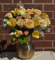 Yellow roses, tulips, carnations and ranunculuses arranged in a long golden vase, with a small bunny toy resting on top of the bouquet. The elegant vase complements the vibrant flowers, creating a warm and charming floral display