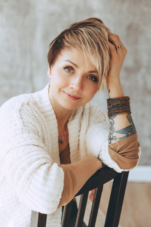 A woman with a short blonde and brown pixie cut poses on a black chair, wearing a white knit cardigan, a brown sleeveless top, and multiple stacked bracelets.