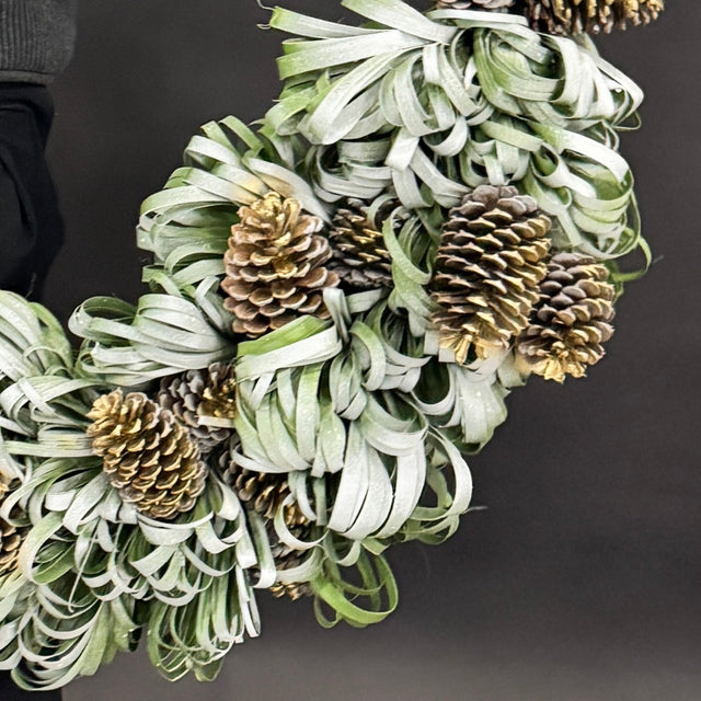 Close-up of snowy palm leaves and golden pinecones on handcrafted holiday wreath.