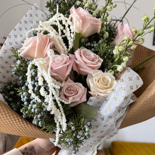 Bouquet with light pink roses, juniper, white delphinium, and hanging amaranthus wrapped in eco-friendly kraft paper.