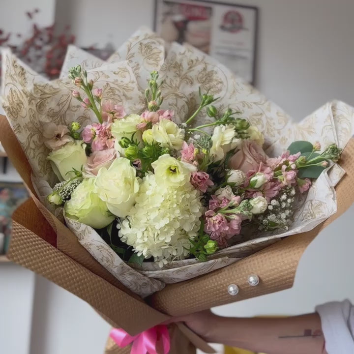 Video showcasing a beautifully arranged bouquet with white hydrangeas, white roses, ranunculus, and light pink stock flowers. The bouquet, elegantly wrapped in white tissue with a gold pattern, is held by the florist