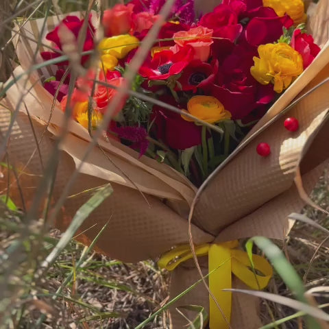 Video of the Juicy Fall bouquet placed in a grassy field, highlighting its vibrant yellow ranunculuses, red roses, orange spray roses, and red anemones against a natural, beautiful backdrop.