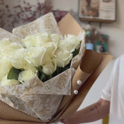 Video of a florist presenting a bouquet of white roses wrapped in kraft paper, adorned with a white ribbon and beads.