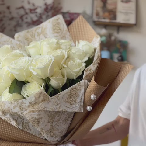 Video of a florist presenting a bouquet of white roses wrapped in kraft paper, adorned with a white ribbon and beads.