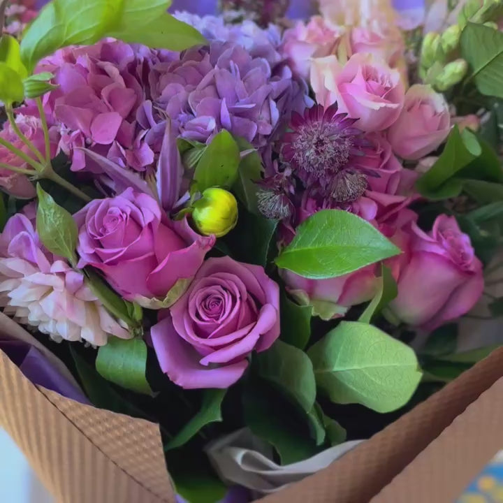 Delicate bouquet of lavender and pink flowers, including hydrangea and snapdragons, wrapped in kraft paper, giving it a warm, rustic appeal.