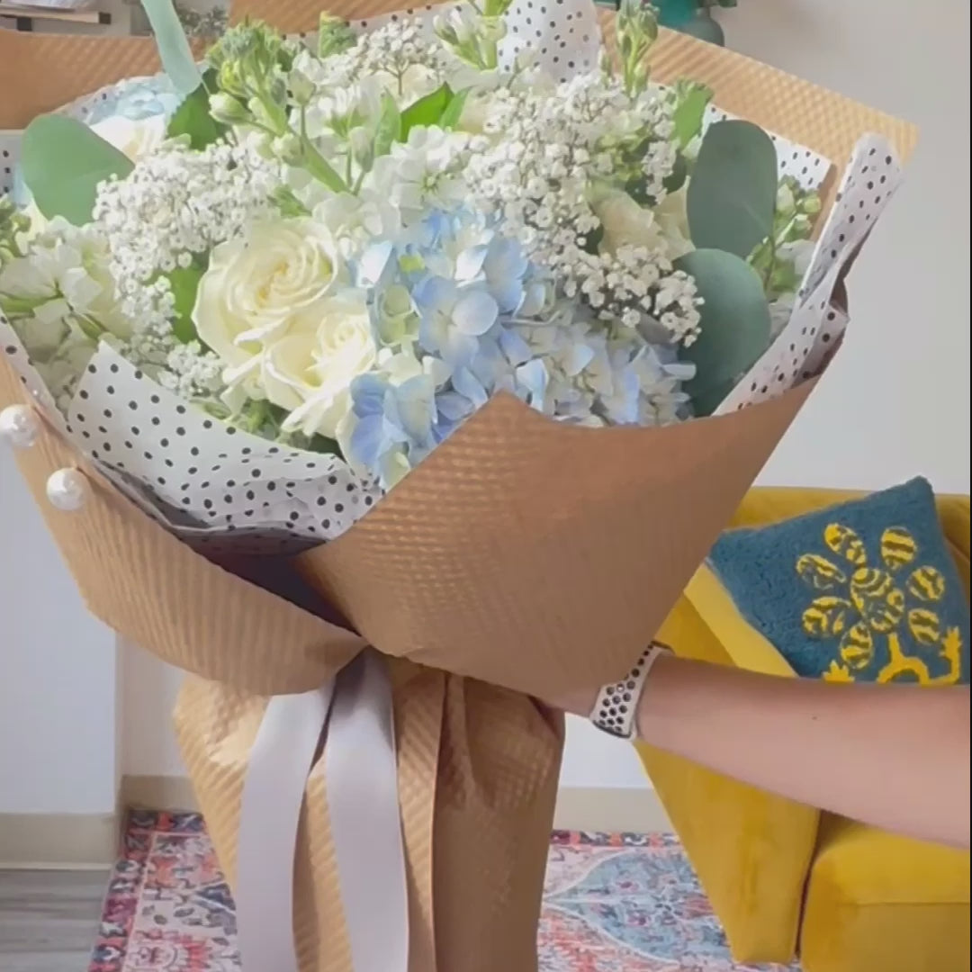 Video of a florist showcasing a bouquet featuring blue hydrangeas, white roses, baby's breath, and stock flowers, highlighting its elegant arrangement