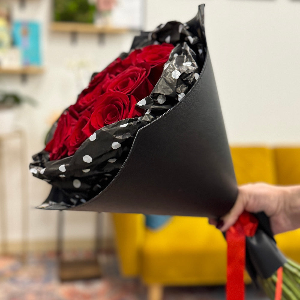 Bouquet of red roses in a black paper cone with polka dots, held by a person in an indoor setting.
