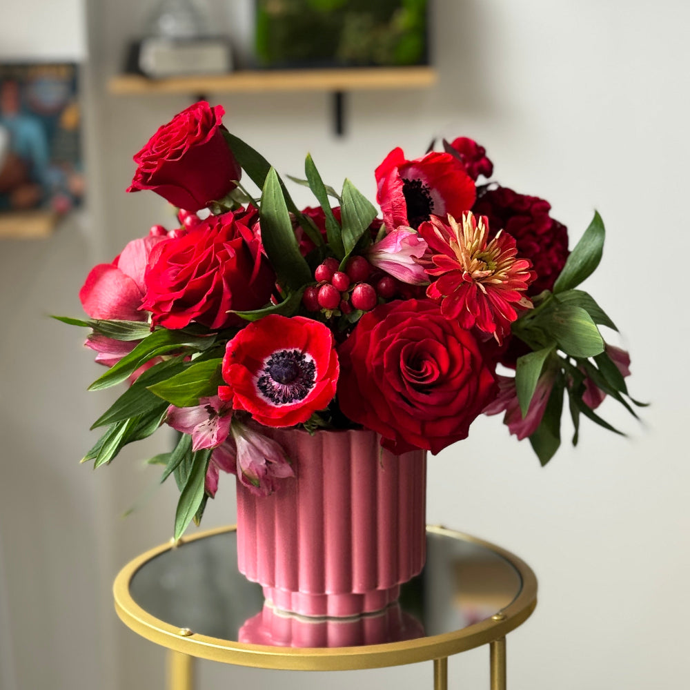 Designer floral centerpiece with rich red blooms, textured hypericum berries, and elegant seasonal flowers in a pink vase.