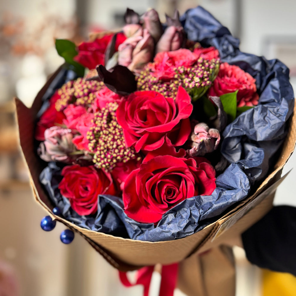 A warm, romantic Rustic Flame bouquet featuring parrot tulips, classic tulips, red roses, spray roses, yarrow, and ruscus in navy-blue and kraft wrapping.