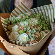 Champagne Dreams bouquet featuring cappuccino-toned roses, creamy white roses, and airy Queen Anne's lace, beautifully wrapped in striped brown and gold tissue paper, with a rustic kraft paper layer. Held gracefully by the florist, the soft neutral tones create an elegant, romantic look.