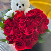 Bouquet of red roses with a white teddy bear holding a heart-shaped card on a blurred background.