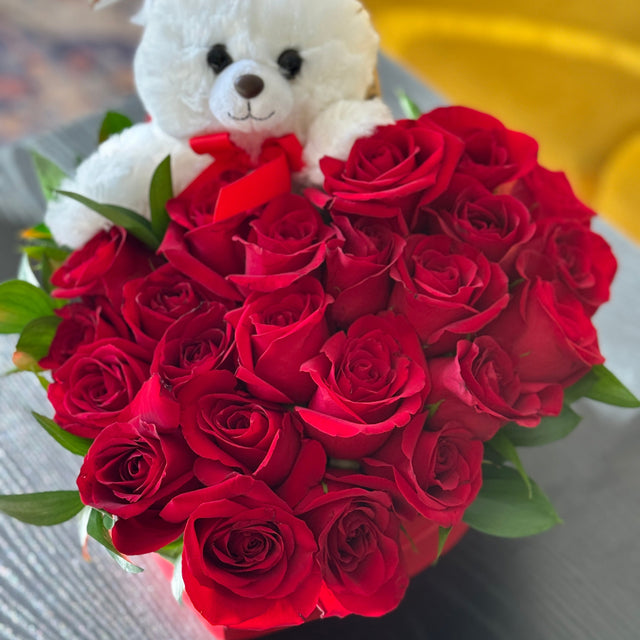 Bouquet of red roses with a white teddy bear holding a heart-shaped card on a blurred background.