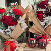 Two bouquets of red roses and greenery in brown paper with ribbons on a wooden crate, surrounded by festive decorations.